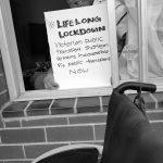 Man sat at a window holding a #LifelongLockdown sign, in the foreground is a wheelchair