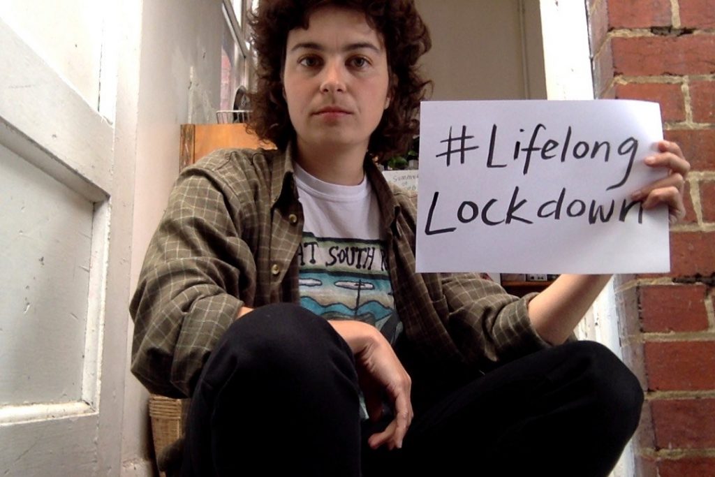 A person sat on a doorstep holding a sign saying #LifelongLockdown