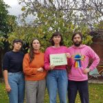 Four people standing in a backyard a green leafy tree is behind them. The third person, a woman with a brown fringe is holding a sign that says #LifelongLockdown