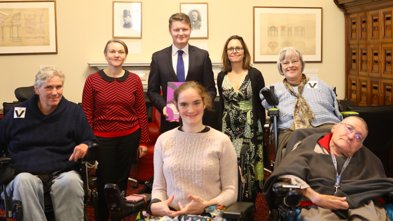 Seven people in an officer of the Victorian Parliament. L-R: DRC Secretary Martin Leckey, a man with wavy grey hair and a blue jumper in a wheelchair; DRC Executive Officer Kerri Cassidy, a woman with short dark hair and bright blue eyes; Member of the Victorian Parliament and Minister for Public Transport Ben Carroll, a man with a young face and short brown hair wearing a suit; Stella Barton, a young woman with light brown hair in a ponytail, wearing a light pink jumper and sitting in a wheelchair; DRC Campaigns Officer Ally Scott, a woman with shoulder-length brown hair and dark-rimmed glasses wearing a black and green wrap dress; DRC Chairperson Mary Henley-Collopy, a woman with shoulder length grey hair and a fringe. She wears clear rimmed glasses and is in a wheelchair; DRC CoM Member Frank Hall-Bentick, a man with close shaved hair, light glasses and a grey jumper sitting in a wheelchair.