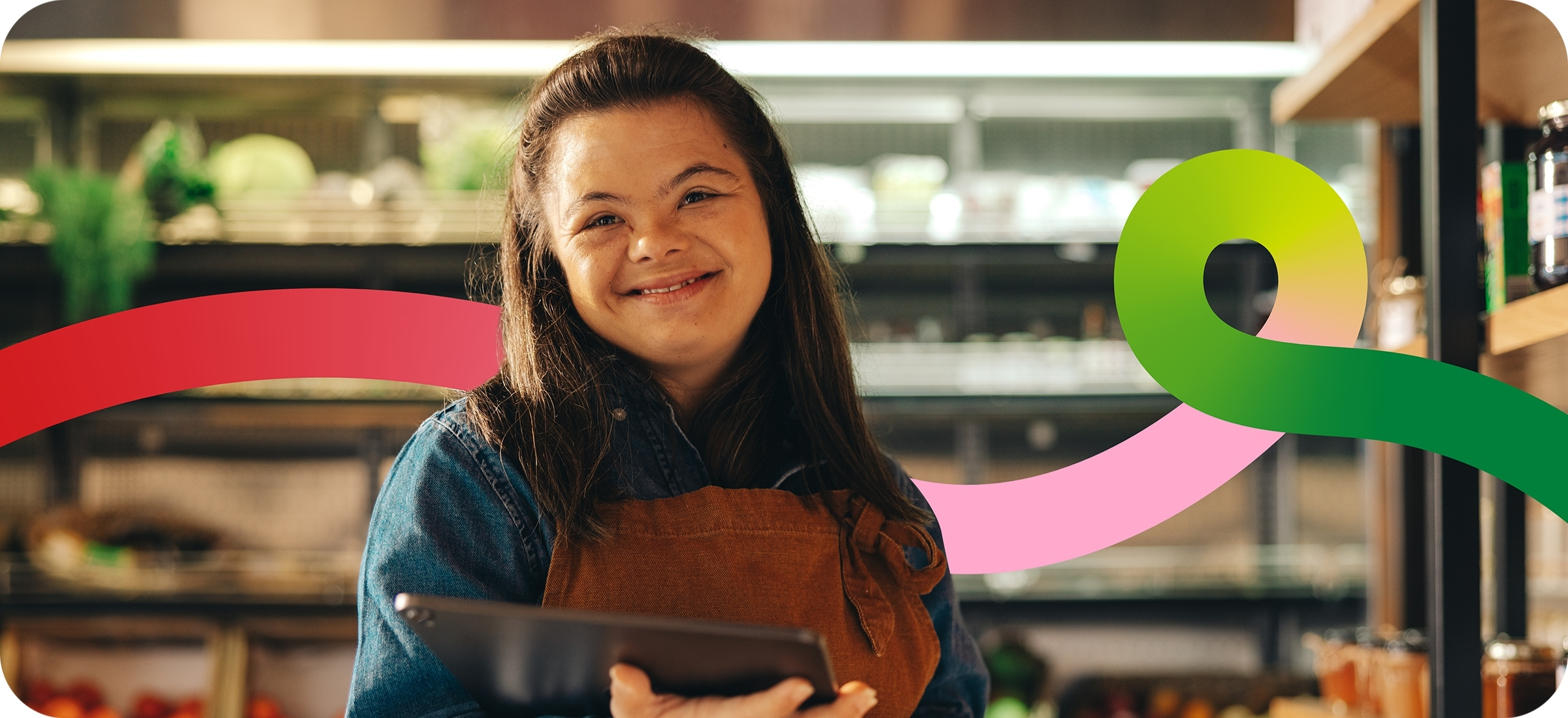 Photo of a person with Downs syndrome who has light skin and long dark hair. They're wearing a brown smock, with a blue denim jacket, and are smiling towards the camera, holding an iPad close to their body. Behind them is a blurry grocery store, with swirls of red and green to match the DRC's logo.