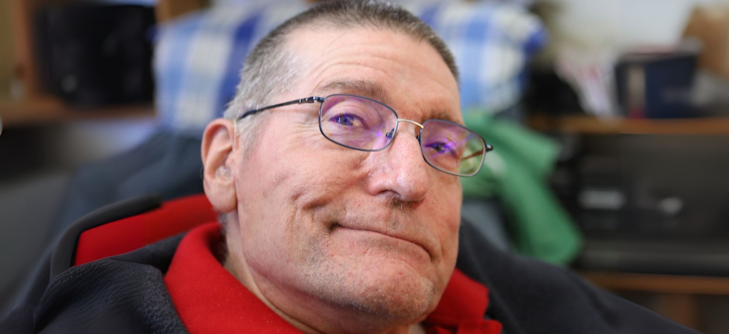 Photograph of Frank Bentick, an older white man with a short grey buzz-cut wearing wire-rimmed glasses and a red polo shirt under a dark jacket. The left-side of his mouth is quirked up in a smile.