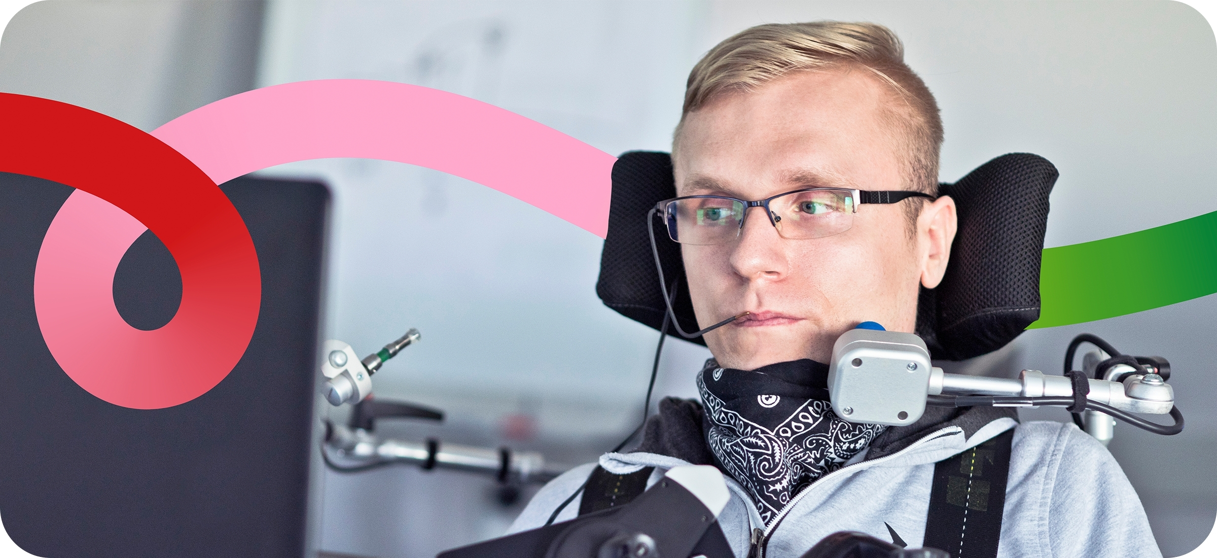 Phot of a person with light skin and short blonde hair, wearing black-rimmed glasses, and sitting in an electric wheelchair with a headrest and other mental assistive technology by their face. They are looking at a computer screen in front of them, with an office setting in the background.