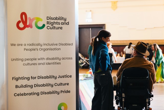 On the right there are two people facing away from the camera, one of them is standing while the other is sitting in an electric wheelchair. On the left of the image is a banner which reads: DRC Disability Rights and Culture We are a radically inclusive Disabled People's Organisation Uniting people with disability across cultures and identities Fighting for Disability Justice Building Disability Culture Celebrating Disability Pride