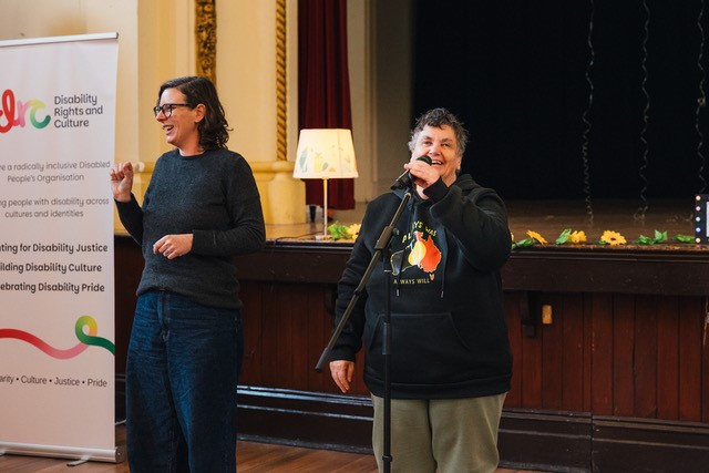 A person standing at a microphone wearing a dark jumper with the aboriginal flag in the shape of so-called Australia on it. There is an AUSLAN interpreter next to them