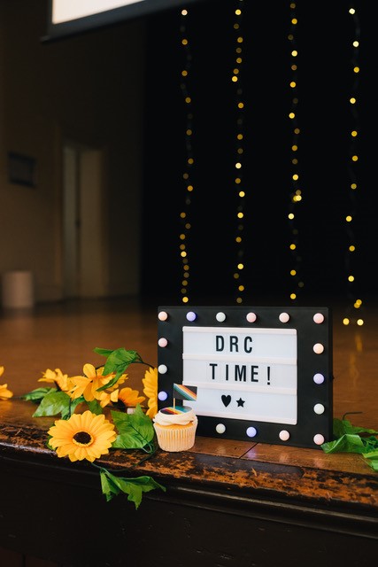 A small screen on a stage that says "DRC TIME!" with a cupcake in front of it and some plastic sunflowers next to it