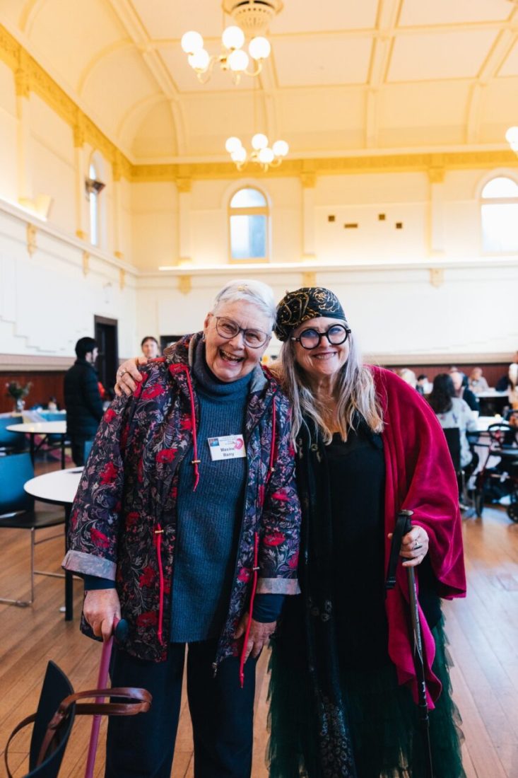 Two people standing next to each other and smiling. One of them has their arm around the other one. Both of them are holding walking sticks