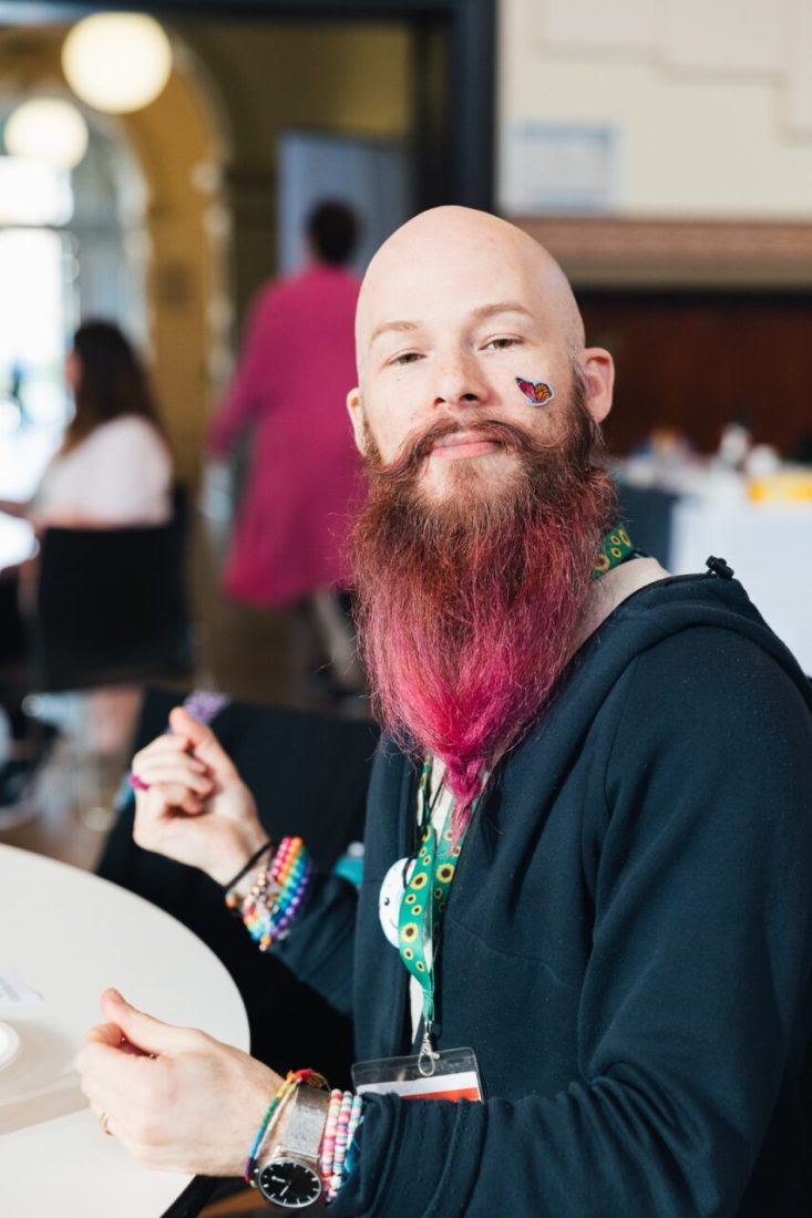 A person looking at the camera. They are bald with a long brown and pink beard. They are wearing a sunflower lanyard and have a butterfly sticker on their face