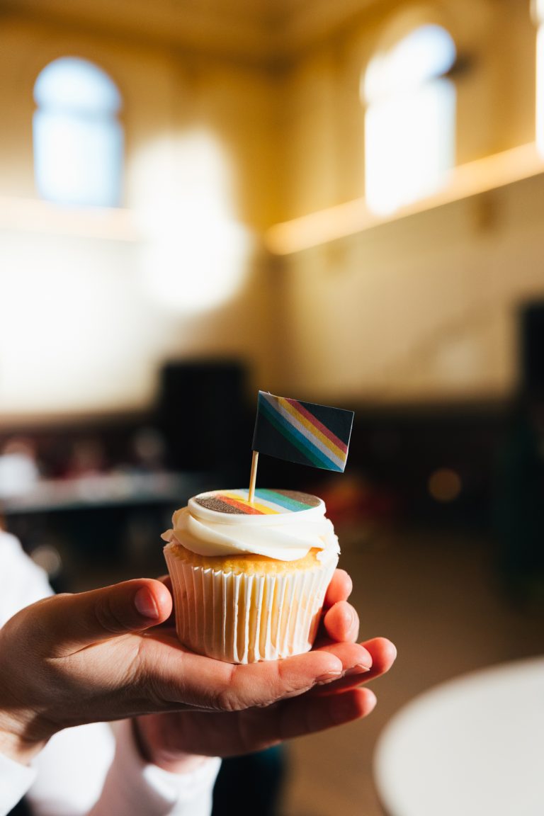 a hand holding a cupcake with an image of a disability pride flag on it. There is also a miniature pride flag stuck in the top