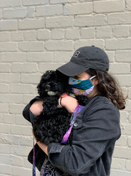 Kelly, a white woman, in 2021 wearing a patterned cloth face mask, a black cap with the Aboriginal flag outlined and holding her sweet lockdown puppy Augie who is a black cavoodle