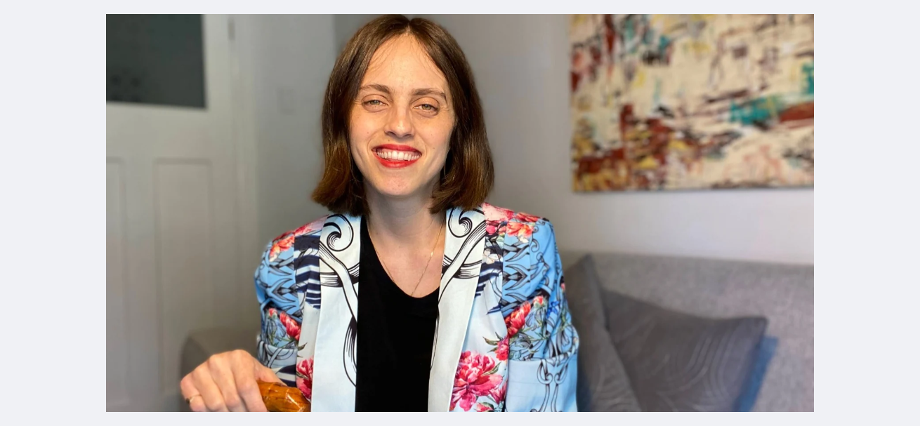 Liel is a femme person with short brown hair and light skin. They are smiling, with bright red lipstick and a colourful blue and white jacket, with a black top underneath.