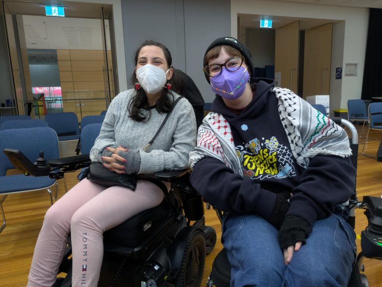 Is and Laura, two writers and wheelchair users, sit next to eachother, both wearing masks for COVID safety.