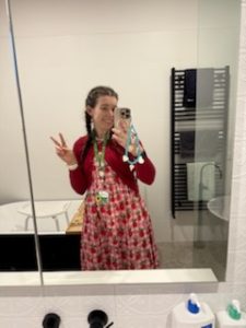 Saffron has tanned skin and medium brown hair. She's smiling in the mirror, taking a selfie, with her hands in the peace sign. She's wearing a cute red dress and jacket, and the hidden sunflower lanyard.