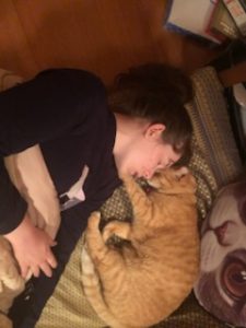 Saffron is a young person with pale skin and dark har, laying face down in bed, looking extremely exhausted and unwell.