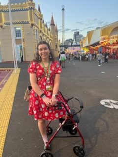 Saffron has tanned skin and long brwn hair, wearing a red dress with white polka dots, posed with her walker. She's standing in a street smiling.