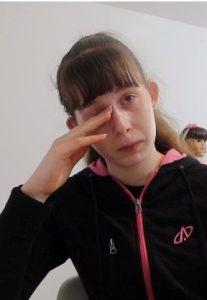 Saffron has light skin and dark brown hair with a fringe. She's rubbing her eyes, as if she has been crying. She looks unwell.