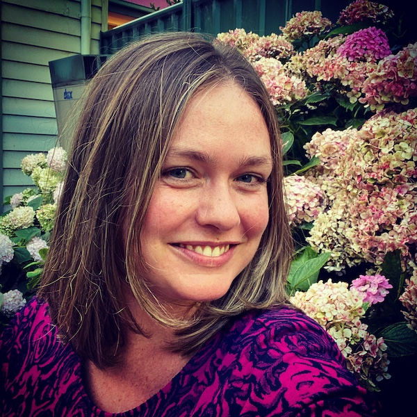 A picture of Emma, a woman with light skin and short dark brown hair with light brown strands. She's wearing a purple dress and smiling in front of a wall of flowers.