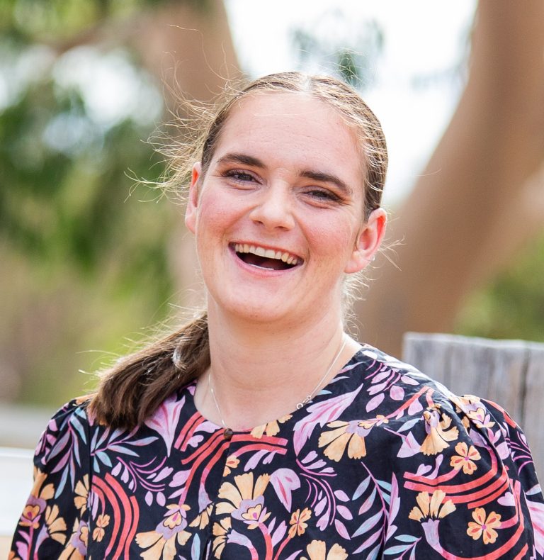 A picture of Stella Barton, a woman with long darkish blonde hair tied up in a pony tail, smiling widely. She wears a dark floral shirt.