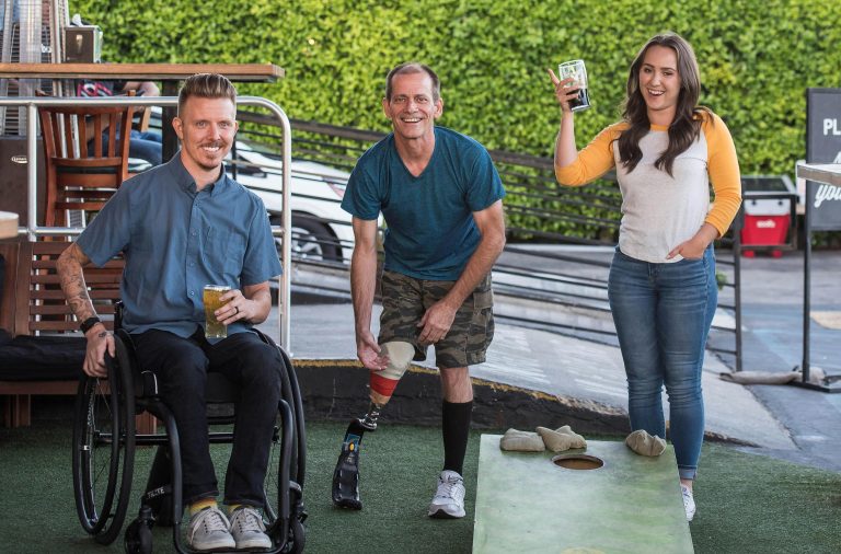 Three people are outside at a barm having fun. One is a masc-looking person sitting in a manual wheelchair holding a beer, one is a masc-looking person standing with a prosthetic leg, and the third is a femme-looking person holding a drink, smiling and standing.