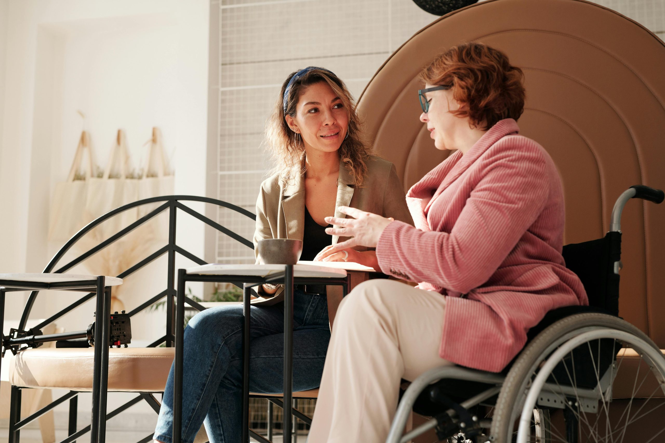 Two people are sitting at a table chatting, with one person sitting in a manual wheelchair. The picture looks warm and inviting.