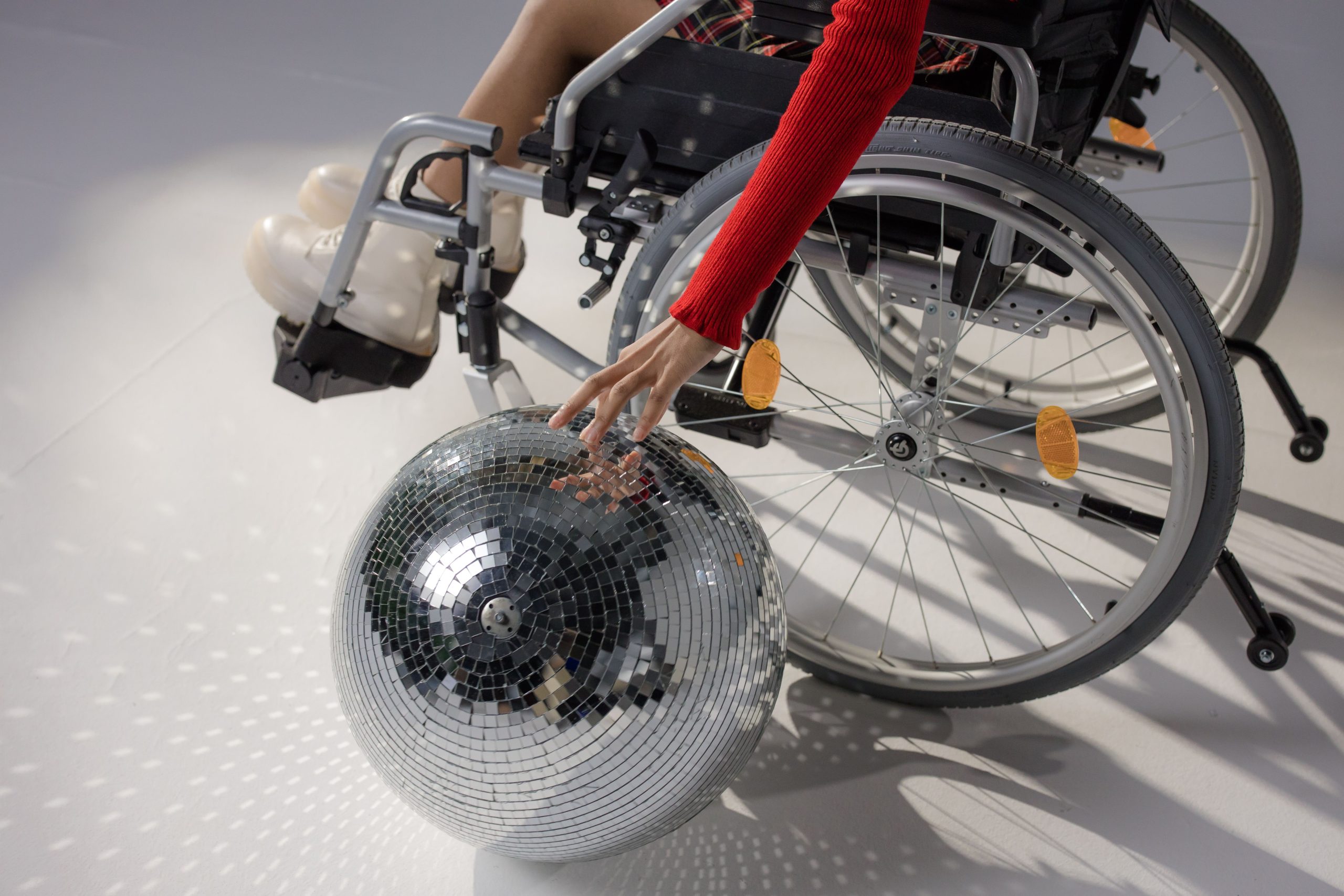 Disability pride means . . . A picture of a manual silver wheelchair with a silver disco ball. A person is sitting in the wheelchair, although you can only see their arm and a red jumper, and a hand which is touching the disco ball.