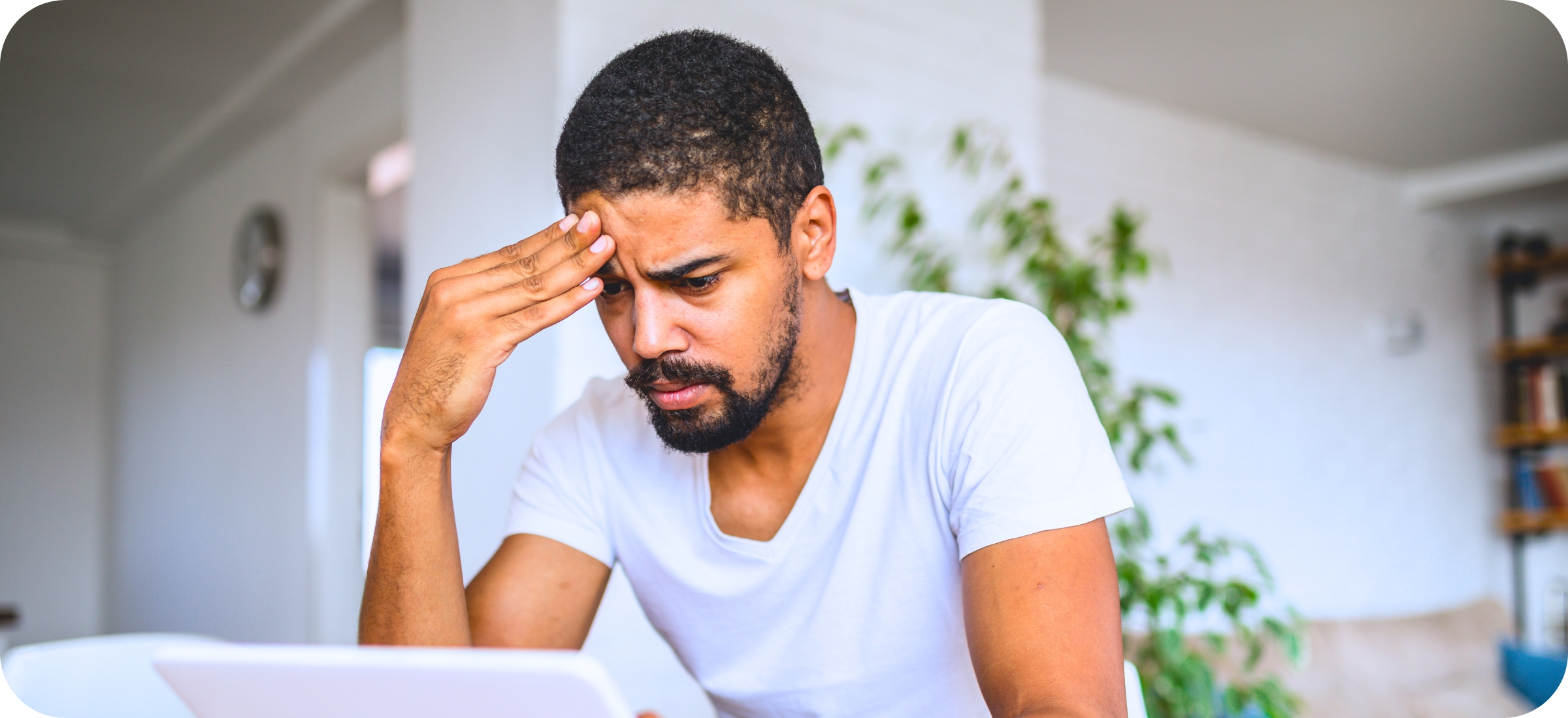 A young man with brown skin and short cropped, black hair and beard, wearing a white tee-shirt. He is leaning forward, head in hand, very worried as he looks at a white tablet.