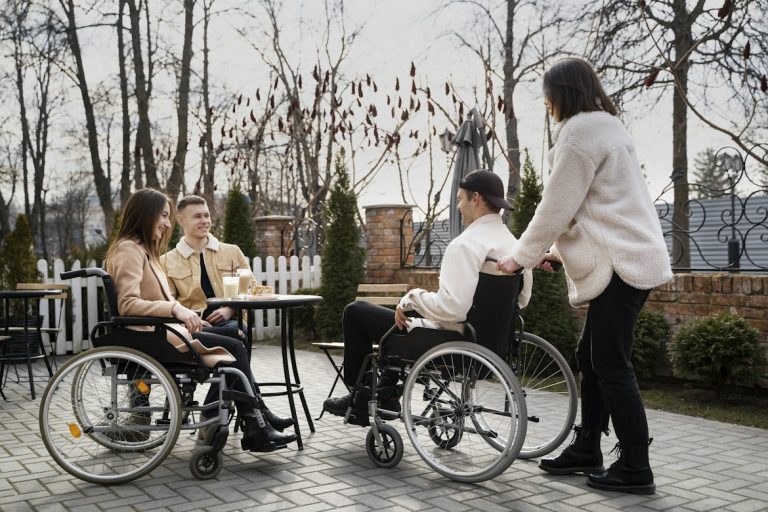 Three people sitting around a table outside, and one person standing. Two of the people sitting are in wheelchairs.